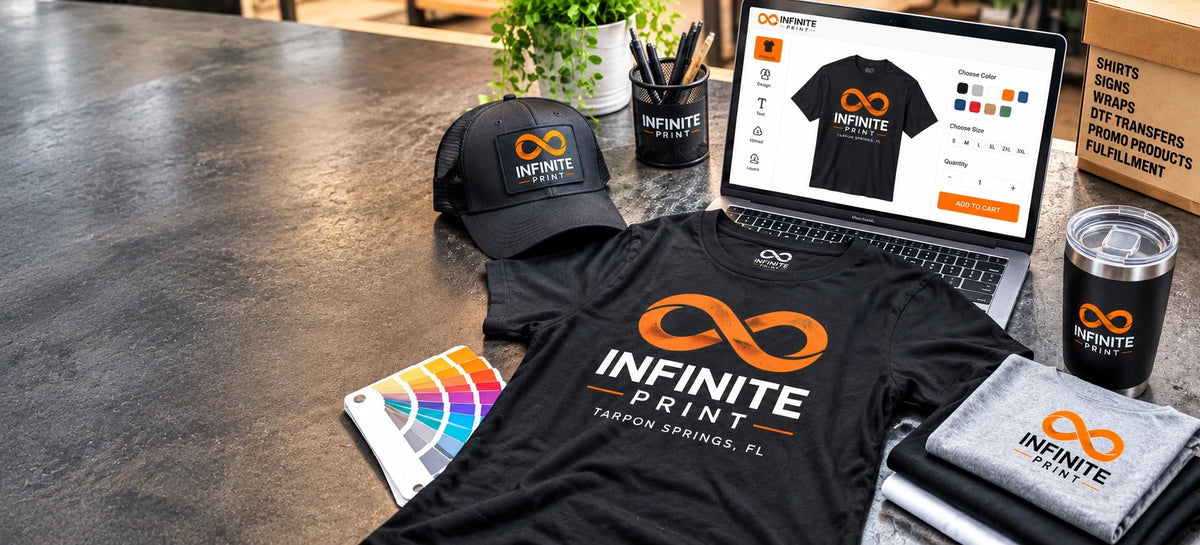 Infinite Print custom apparel on a workbench: a black branded t-shirt, hat, tumbler and folded shirts beside a laptop showing the online shirt designer